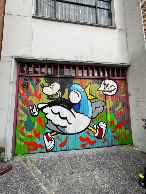 A colorful street art mural painted on a garage door depicting a cartoon duck-billed character running in red sneakers, set against a vibrant red, green, and orange background.