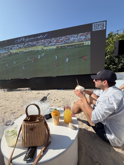 A man enjoys drinks at a beach setup while watching a World Cup match on a large outdoor screen branded with "Polymarket." The scene reflects a luxurious "find your beach" experience for watching the tournament, blending sports viewing with a stylish seaside vibe.