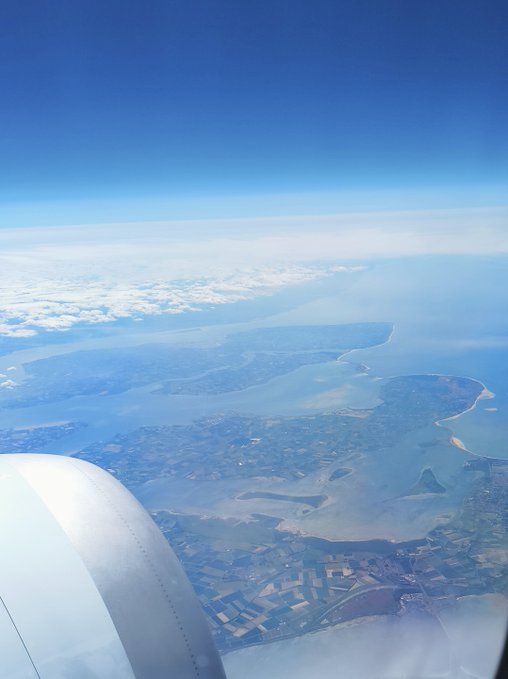 Aerial view from an airplane window showing a coastline with a bay, land masses, and scattered clouds above.