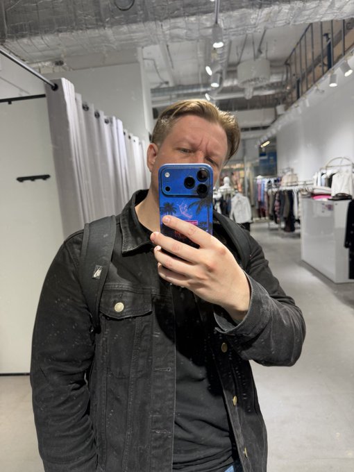 A man with short brown hair wearing a dark denim jacket takes a mirror selfie in what appears to be a clothing store, using a phone with a blue tropical/palm tree case.