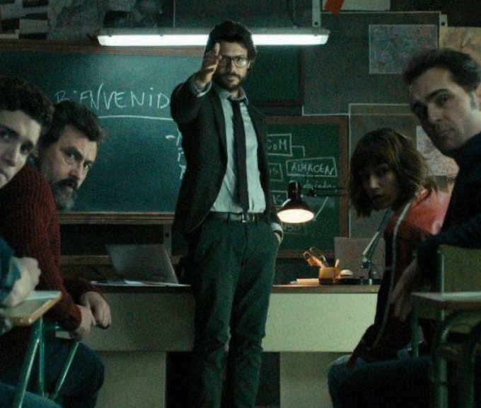 A man in glasses and a suit points forward in a classroom with a chalkboard reading "BIENVENIDOS," surrounded by attentive colleagues, evoking a "professor explains" meme often used to break down complex topics—like how $KAS's kHeavyHash proof-of-work and GHOSTDAG consensus set it apart from typical blockchains.