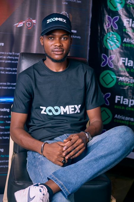 A young man wearing a black 'ZOOMEX' t-shirt and matching cap, seated in a chair at what appears to be a promotional event with 'Flap' and 'Africansi' branded banners in the background.