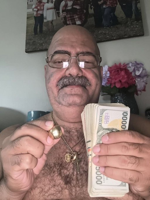 A shirtless older man wearing glasses and gold jewelry holds a thick stack of US $10,000 bills up to the camera.