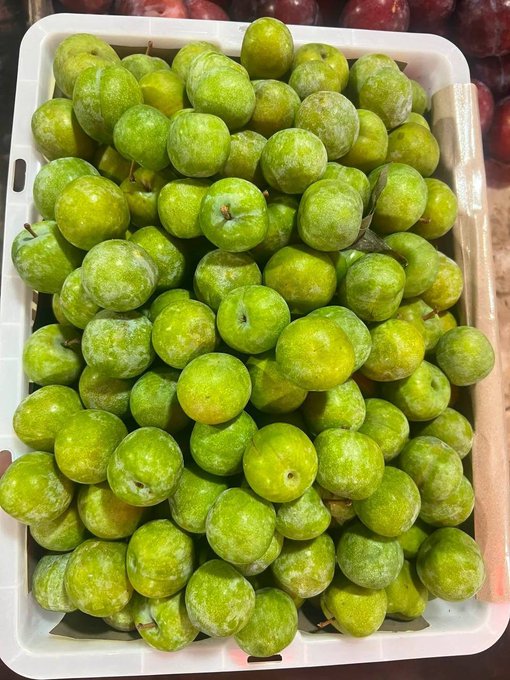 A white crate filled with green plums with a powdery bloom, with red plums visible in the background.