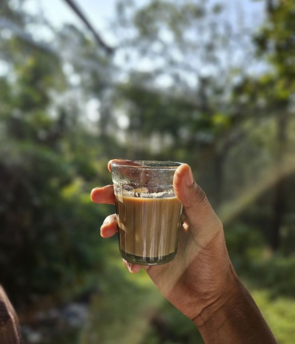 A close-up of a hand holding a glass of milky coffee or tea against a backdrop of blurred green trees and foliage.