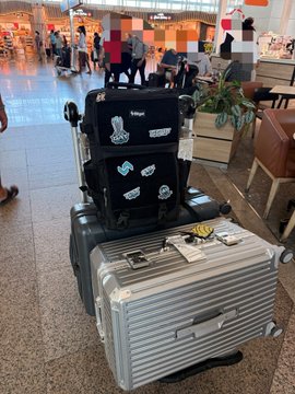A stack of three black Rimowa and silver suitcases sits on a luggage cart in an airport terminal, decorated with travel stickers and ready for the journey home. The image pairs with the caption, symbolizing excitement for the upcoming week with gold and Bitcoin prices on the rise.