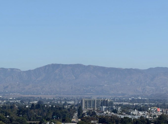 A wide landscape view showing a cityscape with buildings and trees in the foreground and a large mountain range under a clear blue sky in the background.