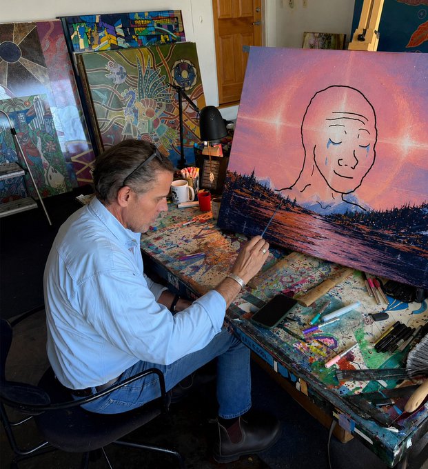 An older man with glasses on his head sits at a paint-splattered desk, working on a large painting of a serene face with a pink, starry background.