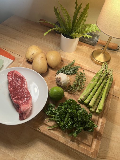 A wooden cutting board displays ingredients including raw steak, potatoes, garlic, lime, asparagus, and fresh herbs, with a potted plant and lamp in the background.