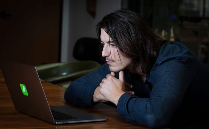 A man with long dark hair leans intently toward a laptop screen displaying a green logo, his hands clasped in deep concentration. The image suggests the focused, solitary effort behind the joke—"someone had to"—implying he took on the unglamorous or thankless task of creating that familiar green symbol.