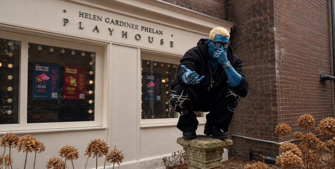 A person in full blue body paint and a black jacket crouches on a stone pedestal while talking on a phone, posed dramatically in front of the "HELEN GARDINER PHELAN PLAYHOUSE" building, with dried hydrangea bushes in the foreground.