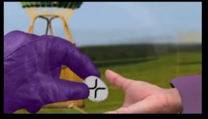 The image shows a hand holding a silver coin that says "XRP" on it. The person is wearing purple gloves and appears to be interacting with someone outside of the frame, possibly at an event or gathering related to Bitcoin or digital currency. In the background, there are two hot air balloons visible in the sky, adding a visually interesting element to the scene. The image suggests that the coin may have been gifted as part of a celebration or special occasion centered around the theme of digital assets and cryptocurrencies.