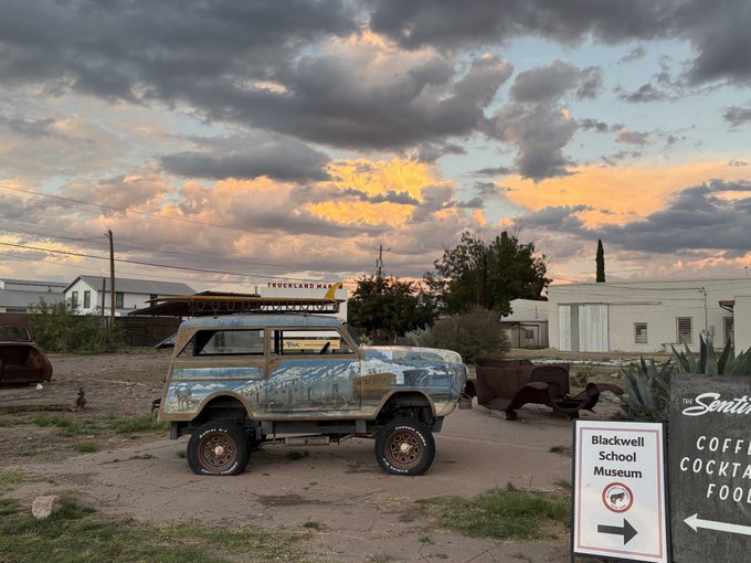 A weathered, lifted vintage station wagon is parked outside the 'Blackwell School Museum,' with a 'TRUCKLAND MALL' sign visible in the distance under a cloudy sunset sky.