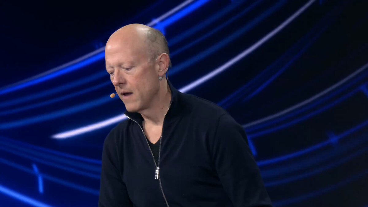 The image features a bald man wearing a black jacket, speaking into a microphone with his hands behind his back. He appears to be delivering a speech or presentation, as indicated by the presence of a computer screen in the background that displays text related to artificial intelligence (AI) and digital identity for AI agents. The caption at the bottom of the image reads "ENS is here in lockstep. Digital identity for AI agents".