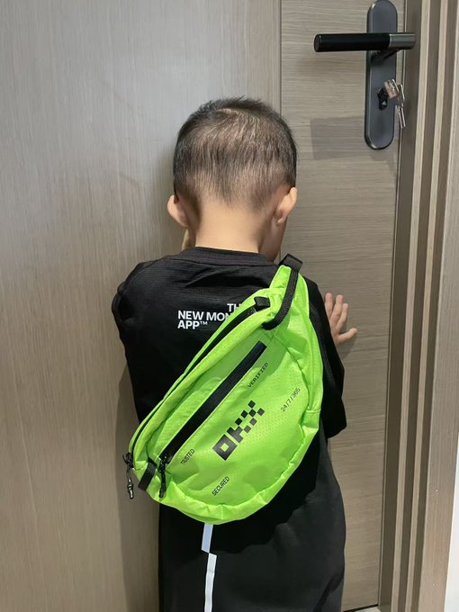 A young boy with a buzz cut stands facing a wooden door, wearing a neon green sling bag and black shirt.