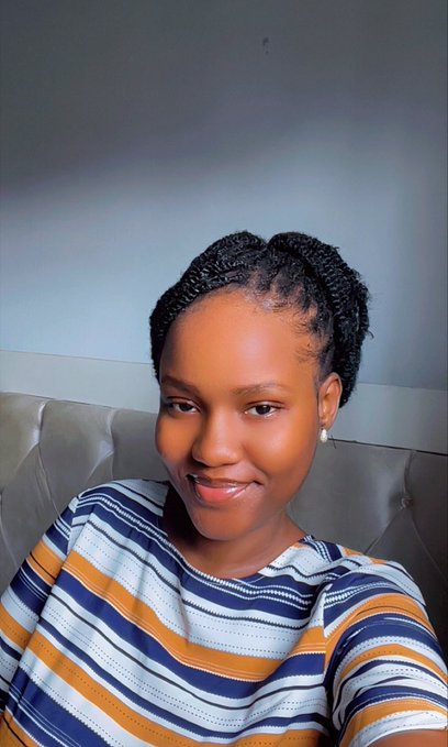 A selfie of a smiling young woman with braided hair wearing a striped top, seated against a gray tufted backdrop.