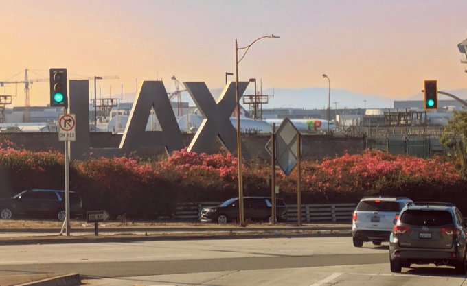 A street scene at dusk features a large, iconic "LAX" sign in the foreground, with cars driving on the road, traffic lights showing green, and airport infrastructure and mountains visible in the background.