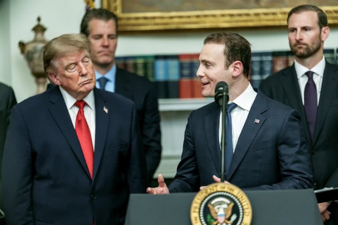 Donald Trump stands beside a younger man in a suit speaking at a podium bearing the Presidential Seal, with two other men standing behind them in an ornate room.