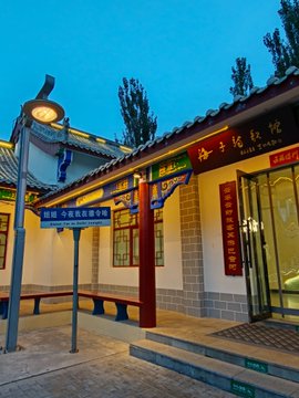 An exterior view of a traditional Chinese building at dusk featuring ornate tiled roofs, decorative wooden brackets, and red banners with gold Chinese characters.