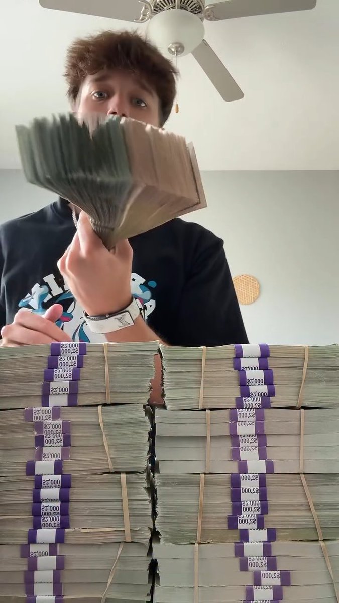 A young man holding up a large fan of cash in front of massive stacks of banded $25,000 bills.