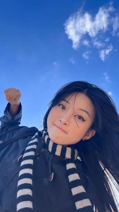 A low-angle selfie of a young woman with long dark hair wearing a striped scarf and dark jacket, raising her fist against a bright blue sky with scattered clouds.