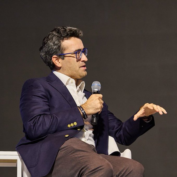 A man with dark hair and glasses, dressed in a blue blazer over a white shirt, speaks into a microphone while gesturing with his other hand during what appears to be a panel discussion or interview.