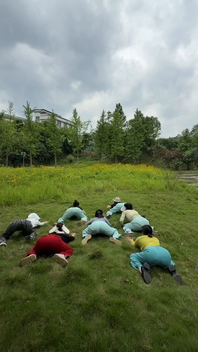 The image features a group of people lying down on grass, possibly taking a break or relaxing during an outdoor activity. The scene includes at least nine individuals scattered across the field, with some closer together while others are more spread out. In the background, there is a house visible behind the group, adding to the overall atmosphere of the image.
