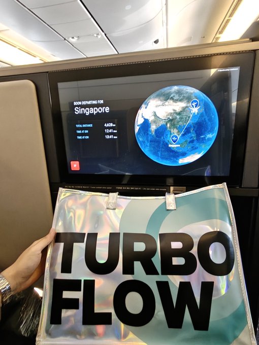 A photo taken inside an airplane cabin showing a person holding a silver bag labeled 'TURBO FLOW' in front of a seatback screen displaying a Singapore Airlines route map for a flight to Singapore, with a total distance of 4,523 miles and a time of 12:41 hours.