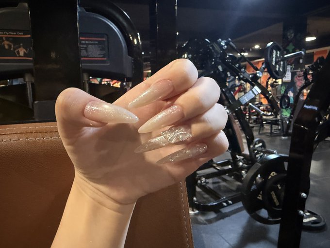 A photo of a hand displaying long, glittery manicured nails inside a gym, with weightlifting equipment visible in the background.