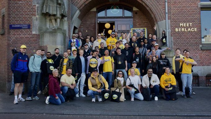 A large group of people pose for a photo in front of a brick building with signs reading 'OUDEMANSSTEEG AMSTERDAM' and 'MEET BERLAGE'.