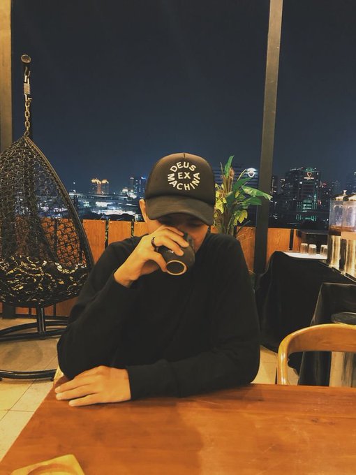 A person wearing a black 'DEUS EX MACHINA' cap and black sweater, drinking from a cup while sitting at a wooden table with a nighttime city skyline in the background.