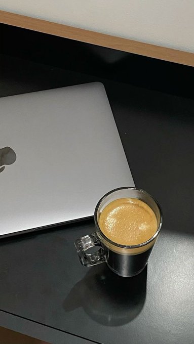 A glass cup of espresso with a thick crema sitting on a dark surface next to a closed silver Apple MacBook laptop.