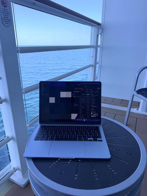 A silver MacBook laptop sits open on a round outdoor table on what appears to be a cruise ship balcony, with the ocean visible through the glass railing.