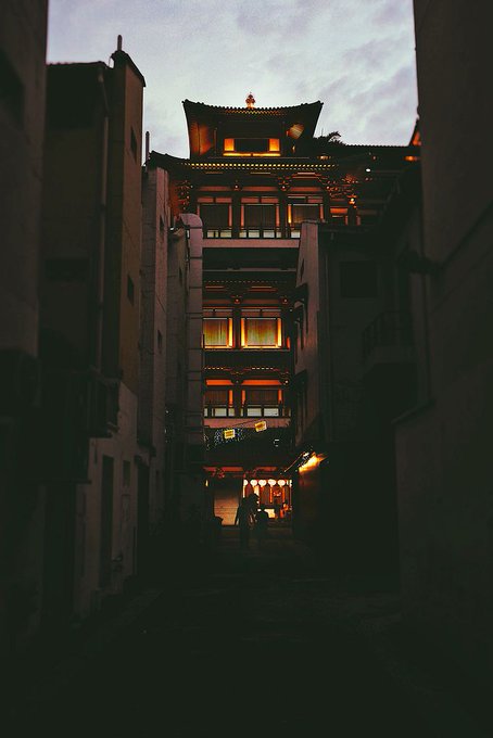 A moody, low-light photograph looking down a narrow alleyway toward a traditional multi-tiered Asian building with a pagoda-style roof, illuminated with warm orange light against a twilight sky.