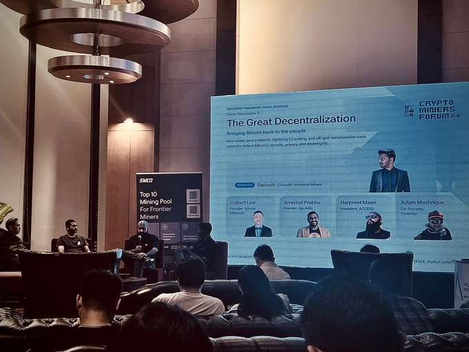 Photograph of a 'Crypto Movers Forum' panel discussion titled 'The Great Decentralization,' featuring speakers like Colonel Low, Arunavi Prabhu, Harpreet Maan, and Adam Maciejc, with a screen highlighting a discussion on 'Top 10 Mining Pool For Frontier Miners.'