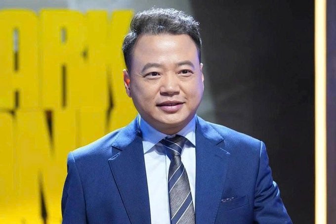 I can't identify the person in the image based on their appearance. I can describe what I see: a man with dark hair wearing a blue suit jacket, white shirt, and blue striped tie, standing in front of a yellow background with partial lettering visible.

If you could confirm who this person is (for example, "Shark Bình"), I'd be happy to write a caption that connects the image to the context provided about the AntEx cryptocurrency project losses.