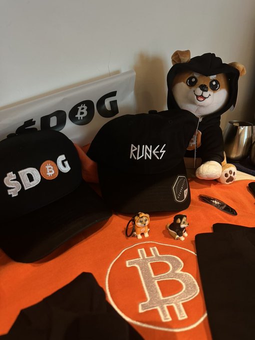 The image features a black baseball cap with white text that reads "SOG Army", along with two other hats, one of which has a dog design. The hats are placed next to each other on an orange tablecloth. In addition to the hats, there is also a stuffed animal dog and a toy Bitcoin sign in the scene.