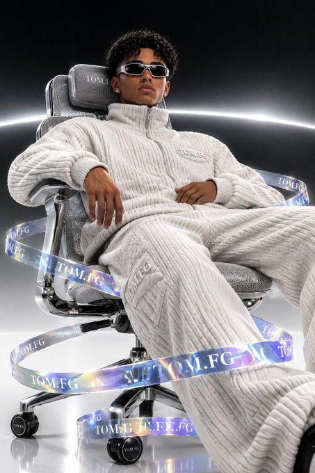 A young man wearing sunglasses and a cream knit outfit reclines in a chair, surrounded by holographic ribbons displaying 'TOM.FG'.