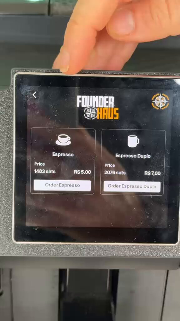 A person's hand pointing at a phone screen showing a price for two coffees including an espresso, cappuccino, double espressos, and regular coffees, all with Bitcoin or Satoshis as payment methods.