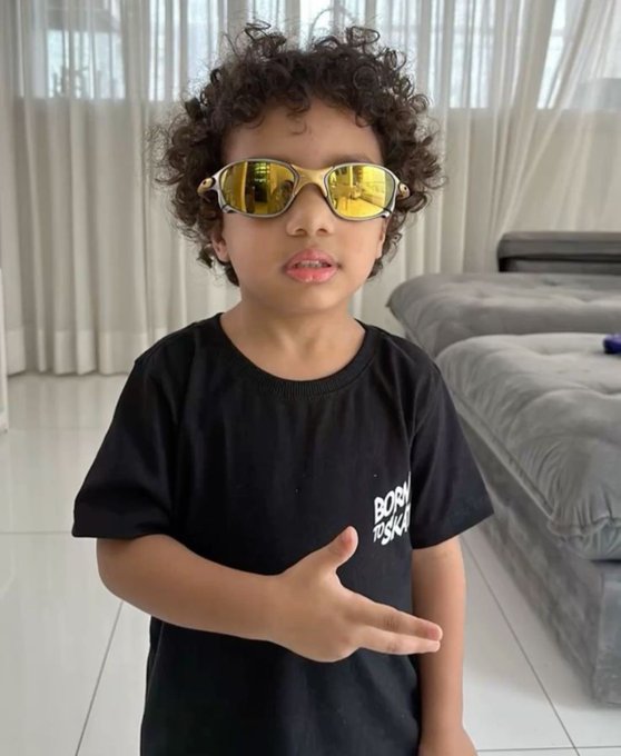 A young child with curly hair wearing reflective yellow sunglasses and a black "BORN TO SHINE" t-shirt, posing indoors.