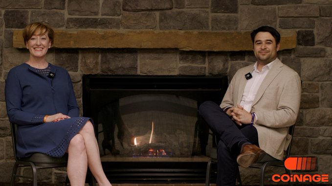 A woman and a man sitting in chairs in front of a stone fireplace, appearing as hosts or guests on the Coinage media channel.