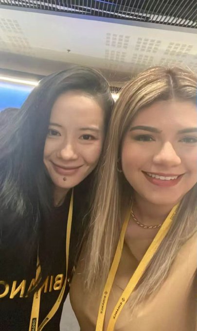 Two young women wearing yellow lanyards pose for a selfie, smiling at the camera with one wearing a black shirt and the other in a white top.