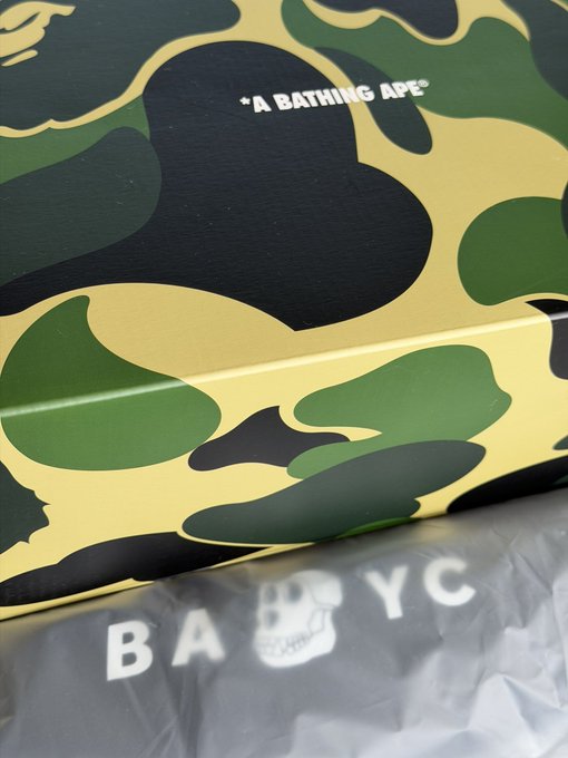 A box featuring "A BATHING APE" branded camo pattern alongside a Bored Ape YC skull logo and "BAYC" lettering on tissue paper, teasing an upcoming Bored Ape Yacht Club unboxing.