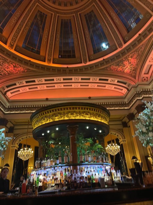 An ornate, domed bar interior with decorative ceiling, chandeliers, and a circular bar displaying numerous bottles of liquor.
