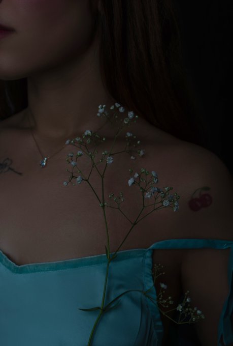 Artistic moody portrait of a woman wearing a turquoise off-shoulder dress and a delicate necklace, holding white baby's breath flowers against her chest, with cherry tattoos visible on her skin.