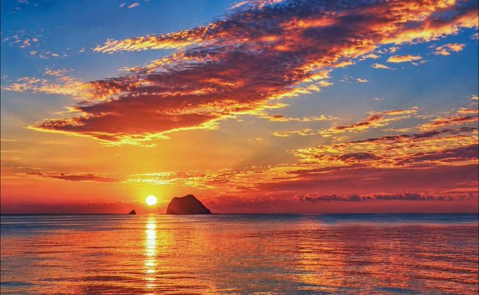 A vibrant sunset over a calm ocean, featuring a bright sun, scattered fiery orange and pink clouds, and two small silhouetted rocks on the horizon.