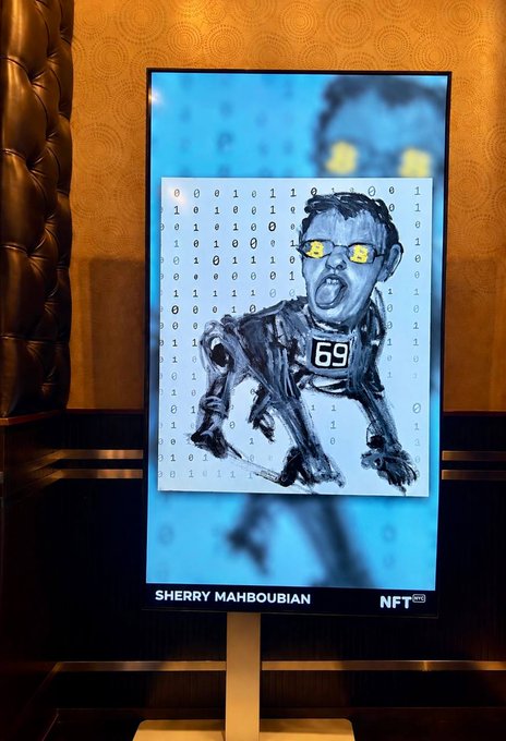 A digital display screen in a venue shows an NFT artwork labeled 'SHERRY MAHBOUBIAN' featuring a stylized portrait of a person with yellow glasses against a background of binary code.