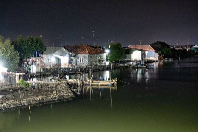 The image shows a nighttime view of a river with several boats floating along its surface. The boats are docked at various points, including one near the shoreline where there's a small house visible. The scene also includes a bridge that spans across the river. In the background, there is a dark sky and trees, creating an atmosphere of tranquility and peacefulness.