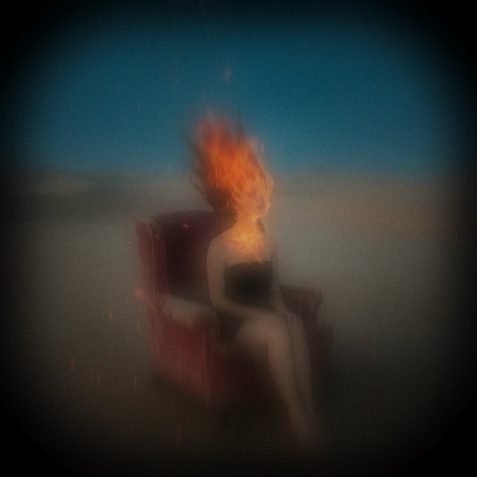 An artistic, blurry image shows a figure sitting in a red armchair with their head engulfed in flames, set against a blue gradient background.