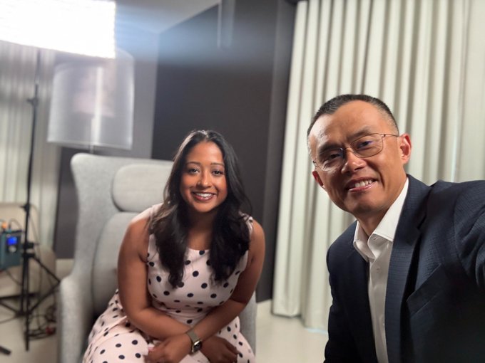 The image shows a man wearing glasses and a suit sitting next to a woman who is also wearing a dress with polka dots, both smiling for the camera. The background features a window with white curtains that allows natural light into the room. The caption of the image reads "One of the most interesting conversations I’ve had this year! Thank you @cz_binance", which suggests that they might be discussing something interesting or noteworthy in their conversation, possibly related to the post on social media.